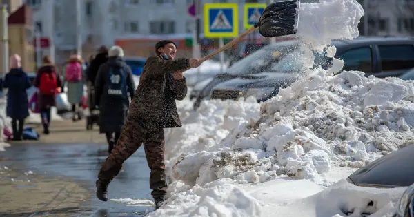 Mann räumt mit einer Schneeschaufel Schnee auf einem belebten Gehweg zwischen parkenden Autos