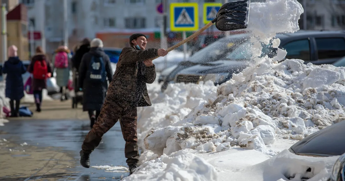 Mann räumt mit einer Schneeschaufel Schnee auf einem belebten Gehweg zwischen parkenden Autos
